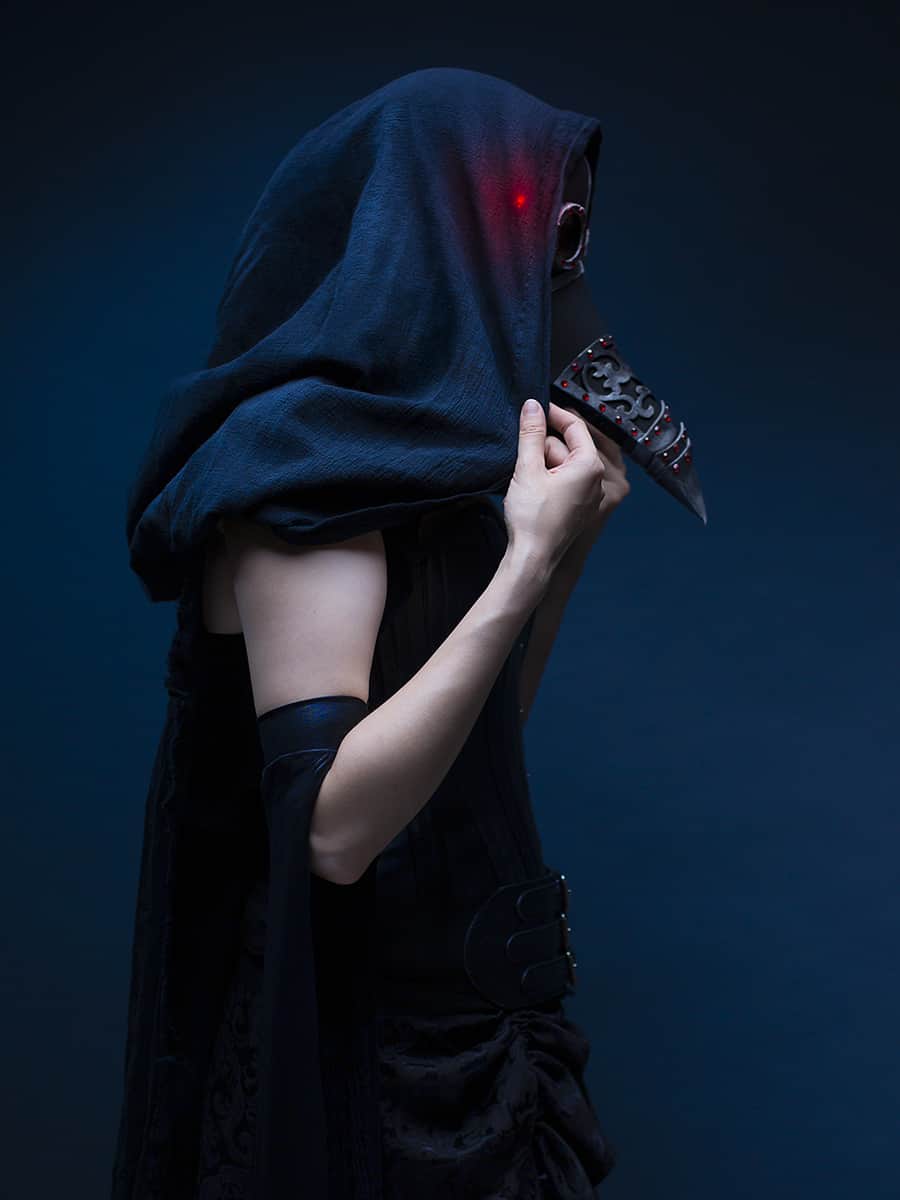 A side profile of Jennifer as the Raven wearing a black cape and a black plague doctor mask with red eyes.