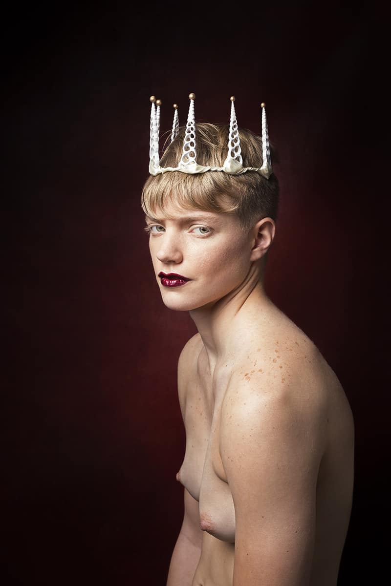 Sarah challenges gender norms with a white shell crown atop short blonde hair, intense blue eyes, red lip, and a strong torso