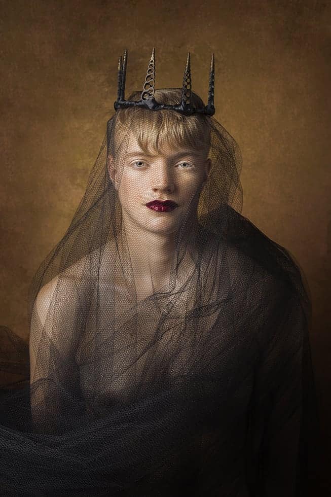 A black spiral shell crown sits atop Sarah's head. Her blonde hair cropped short, blue eyes intense, a bold red lip, and black tulle covering a strong torso